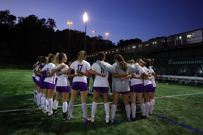 The N.Y.U women’s soccer team wears white and purple jerseys and purple shorts, huddling on the Fauver Stadium in Rochester.