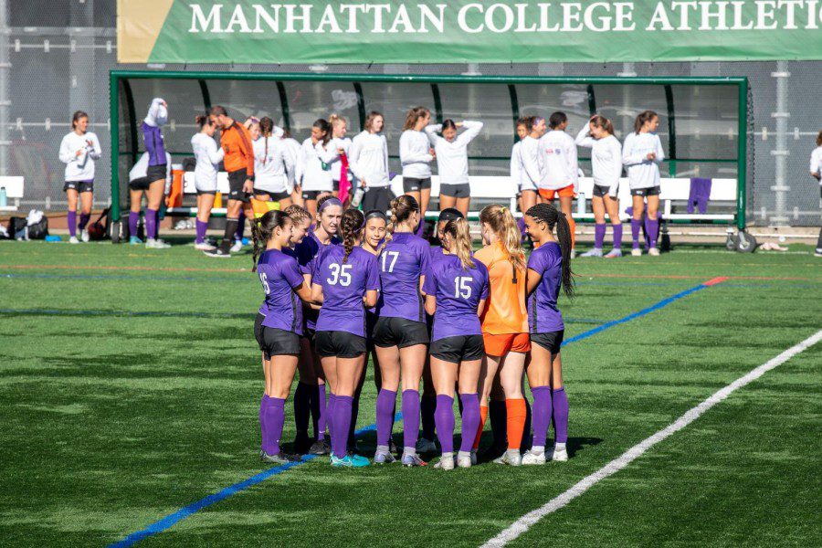 The women’s soccer team huddles in the middle of the field for a discussion under bright sunlight. The team is wearing purple jerseys and socks, and black shorts. The goalie is wearing an orange jersey, shorts and socks. In the background are players from Brandeis University dressed in white and black outfits. The banner in the background reads “MANHATTAN COLLEGE ATHLETICS.”