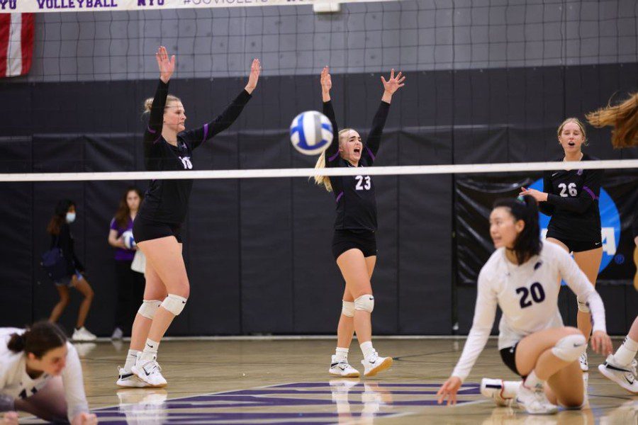 Olivia Lewandowski, Dominique Drust and Sarah Lattan dressed in black jerseys throw their hands up. Volleyball is in mid air.