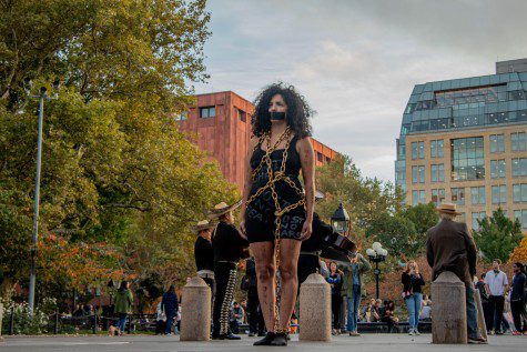 Artist Sofia Saleh wearing a black tank top and shorts with illegible text written on them with white chalk. There is a gold metal chain entangled around her body. Her mouth is taped over with black duct tape. She stands at the north side of Washington Square Park with a group of people behind her.