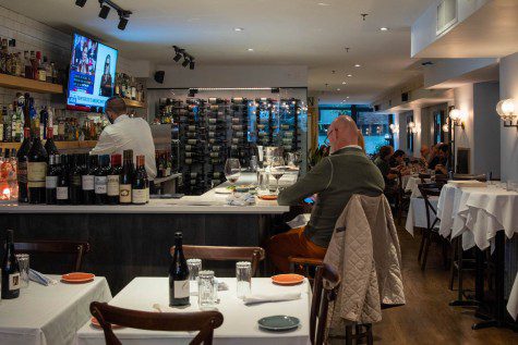 The interior of a restaurant with plates and wine bottles laid across the bar counter. A bartender is working inside the counter with a wall of wine bottles on display behind him and a television screen playing above him.