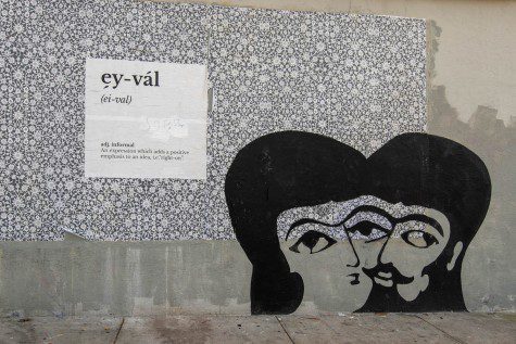 Drawings on a gray wall with flowery white patterns. The phrase “ey-val” is defined as a “a.d.j.informal” and “An expression which adds a positive emphasis to an idea, i.e. “right-on.” Next to the patterns is a cartoonish drawing of two people’s heads merged in the middle.
