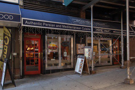 The facade of a restaurant with blue sun shades outside with text “Authentic Persian and Mediterranean Cuisine” written on it in white color. White string lights hang from the awning.