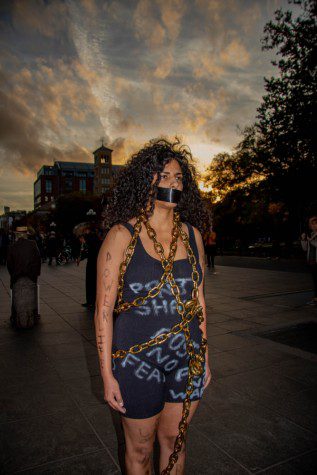 Artist Sofia Saleh wearing a black tank top and shorts with illegible text written on them with white chalk. There is a gold metal chain entangled around her body. Her arms and legs are covered with text “POWER” written with black chalk. Her mouth is taped over with a black duct tape. The sun is setting behind her.