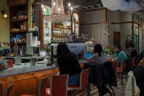 The inside of a cafe with four people sitting at the bar counter. Above them is a chandelier with candle shaped light bulbs.