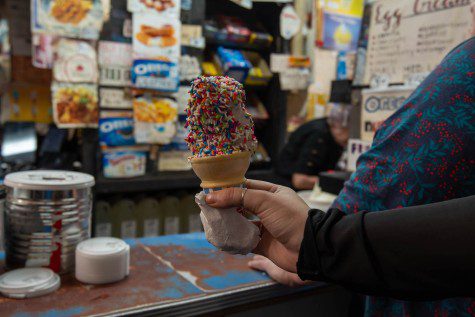 A hand holding a cone of ice cream with candy sprinkles on it. In the background is a counter with cup covers laid on it and rows of snacks hanging on a wall behind the counter.
