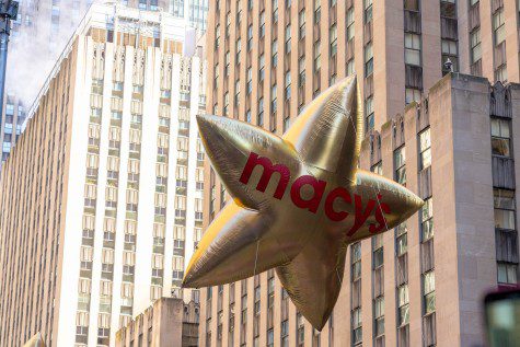 A large, gold, star-shaped balloon floats above the street with skyscrapers in the background. On the balloon there is red text that reads “Macy's.”