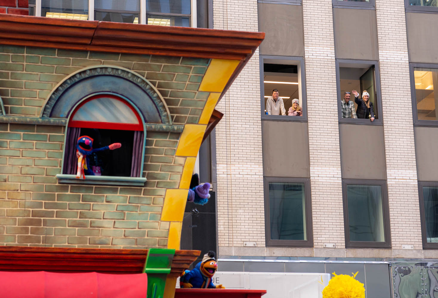 A recreation of a house in Sesame Street featuring Grover, Elmo, and Ernie proceeds on Sixth Avenue. In the background are bystanders looking at the parade through building windows.