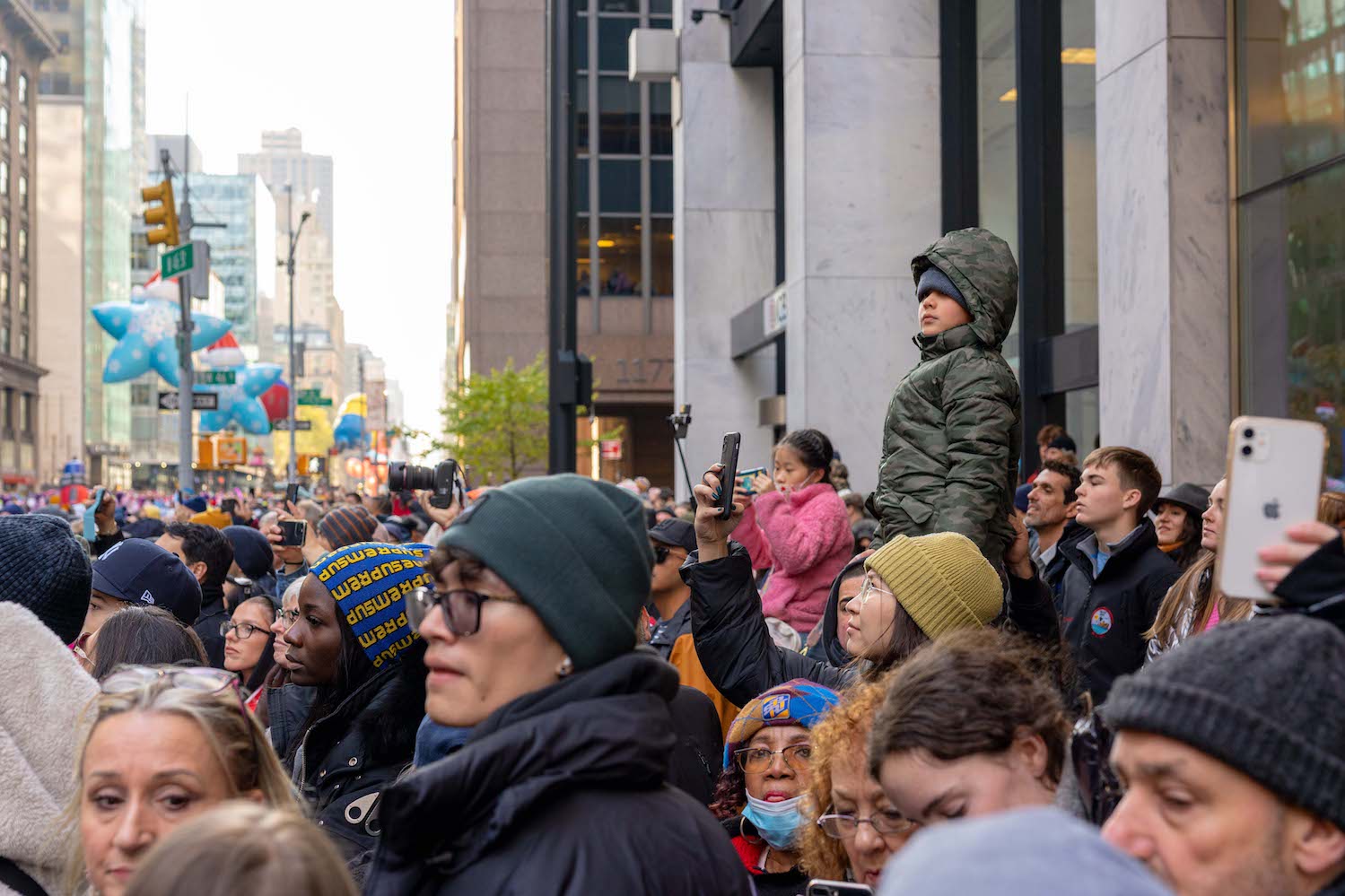 A young boy dressed in a dark green puffer jacket sits atop a man’s shoulders in a crowd of bystanders.