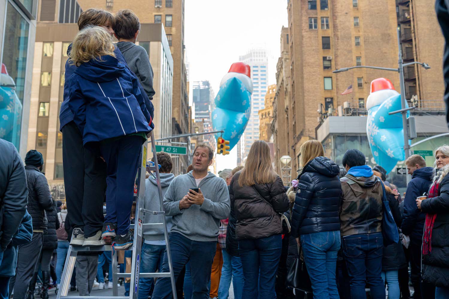 Three kids stand atop a silver ladder to watch the parade behind a line of adults.