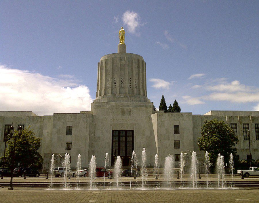 The Oregon State Capitol Building in Salem, Oregon, on a sunny spring afternoon. The exterior facade of the building is constructed of marble, and has been designed in the art deco style. A large dome rises from the center of the building. Atop the dome is a gold statue of a man, called the "Oregon Pioneer."