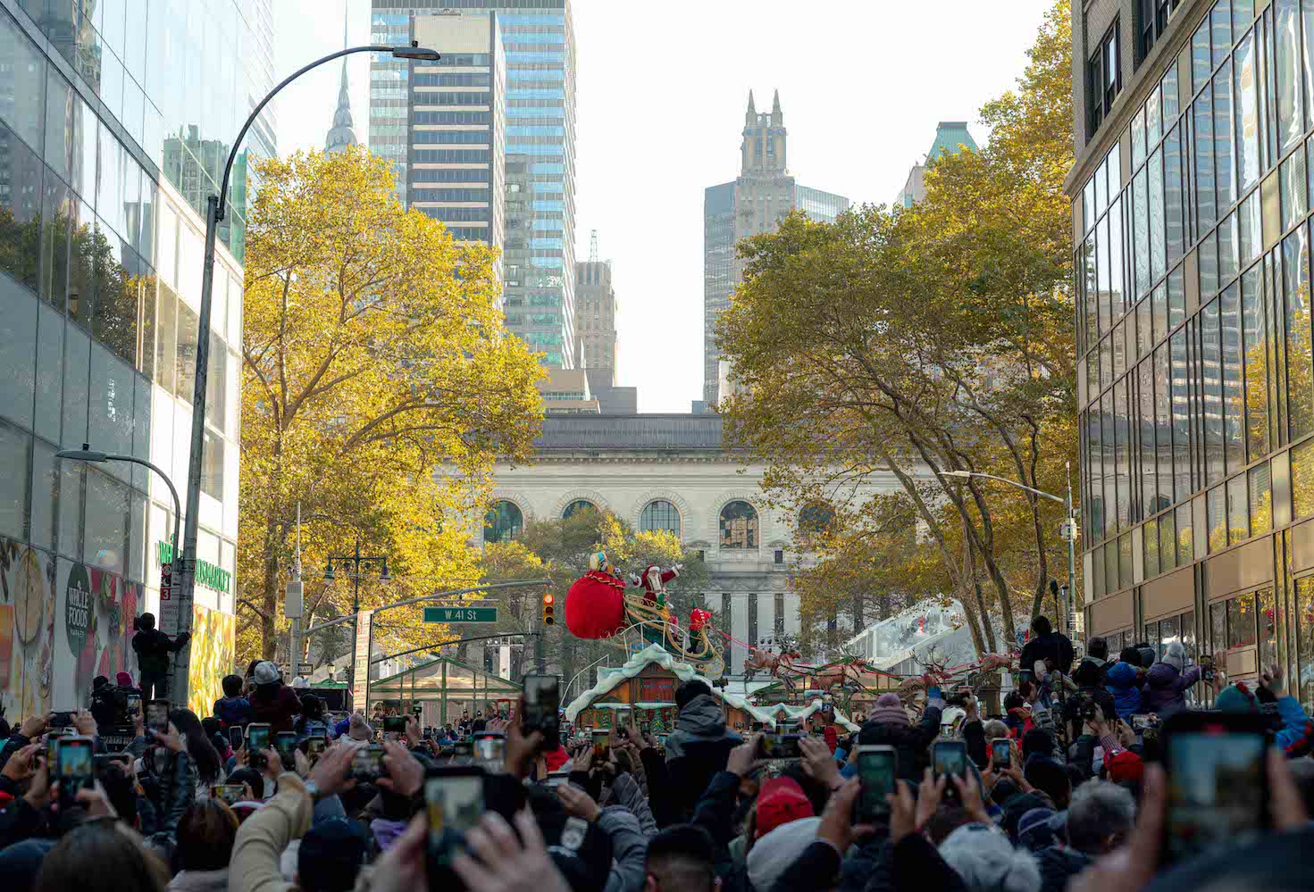 A crowd of bystanders in the foreground watching Santa Claus and his sleigh proceed down Sixth Avenue.