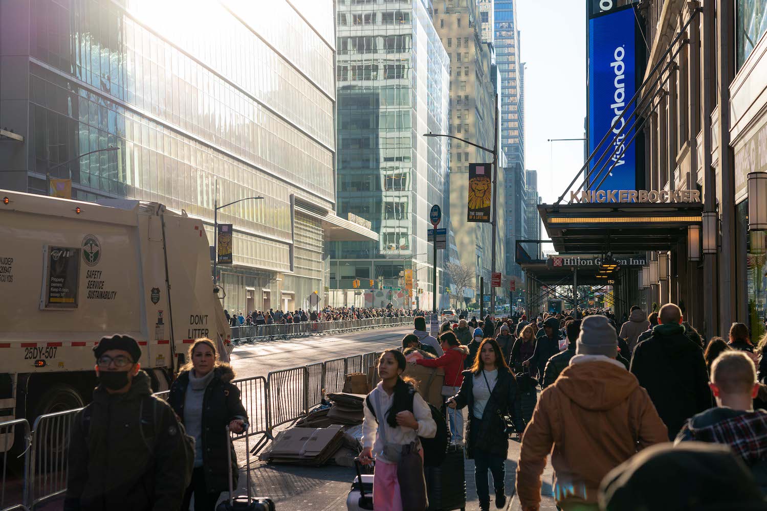 A large crowd of people walk on the sidewalk of Sixth Avenue in Manhattan. The car lanes are fenced off and empty. A garbage truck is parked on the side