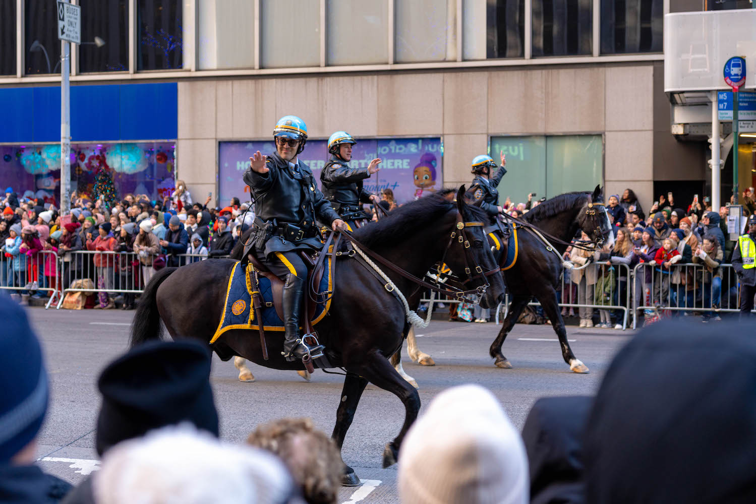 Three police officers wearing blue helmets riding horses down Sixth Avenue in Manhattan. They are waving toward the crowd who gathered behind the fences on the sidewalks