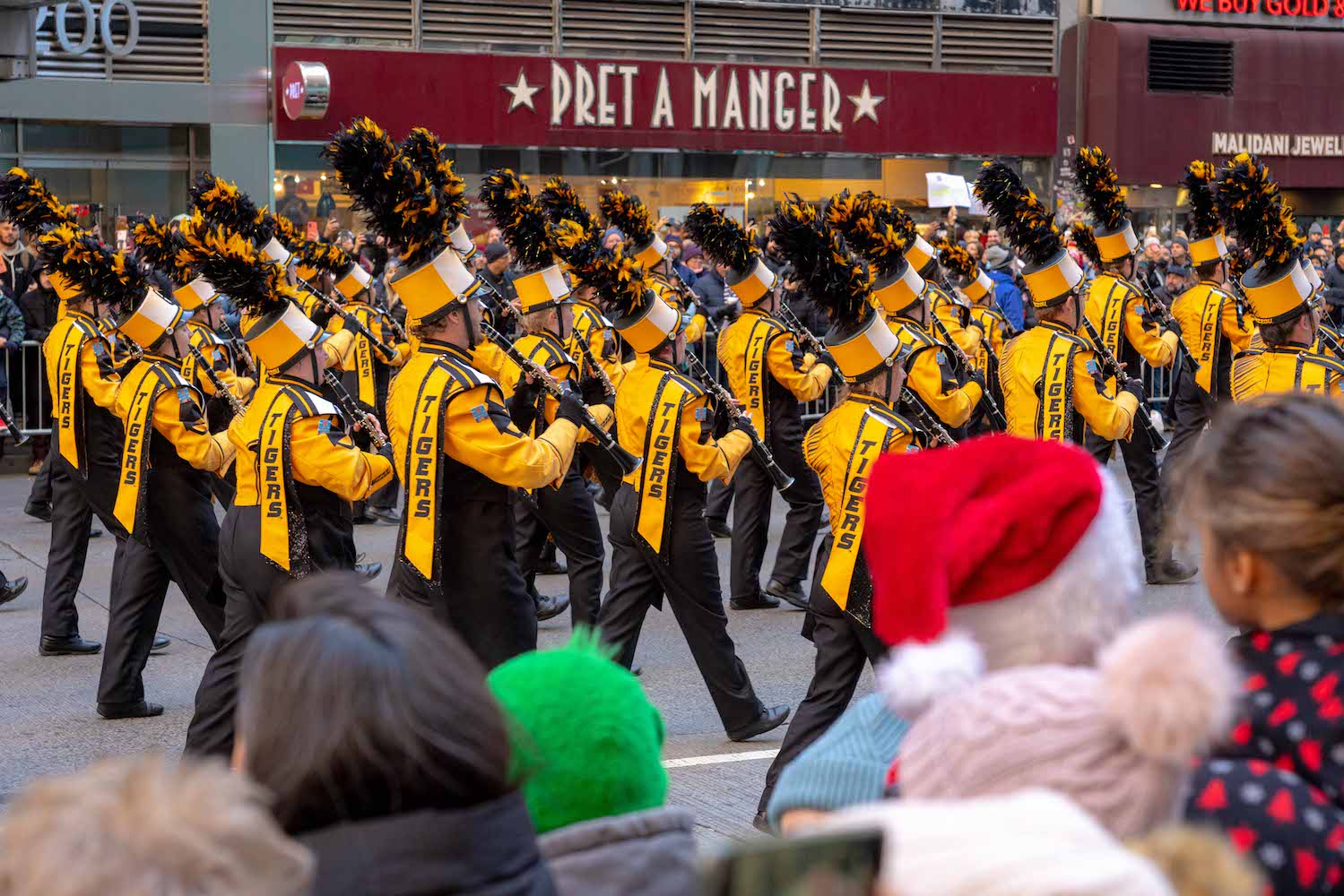 A marching band wearing identical yellow suits, black pants, and feathered hats walking down Sixth Avenue in Manhattan while playing clarinets. In the foreground are crowds watching the parade.