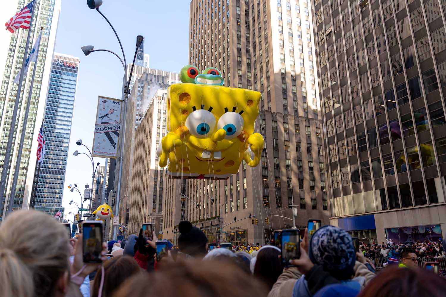 A large balloon in the shape of SpongeBob SquarePants floats above Sixth Avenue in Manhattan while crowds take photos of it with their phones.