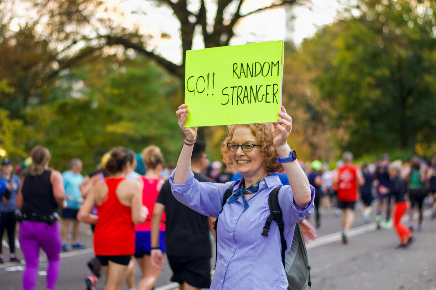 A female wearing a blue long-sleeve shirt and a blue scarf in her neck holds a green poster that reads in black colored font: “Go!! Random Stranger.”