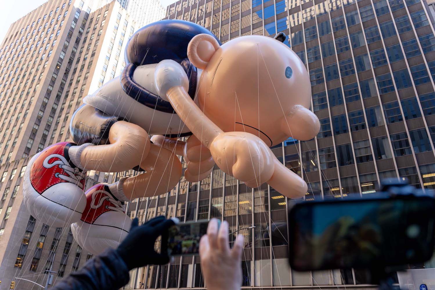 A large balloon of Greg Heffley from Diary Of a Wimpy Kid. He is a young school boy dressed in a white t-shirt, black shorts, and red and white sneakers wearing a black backpack.
