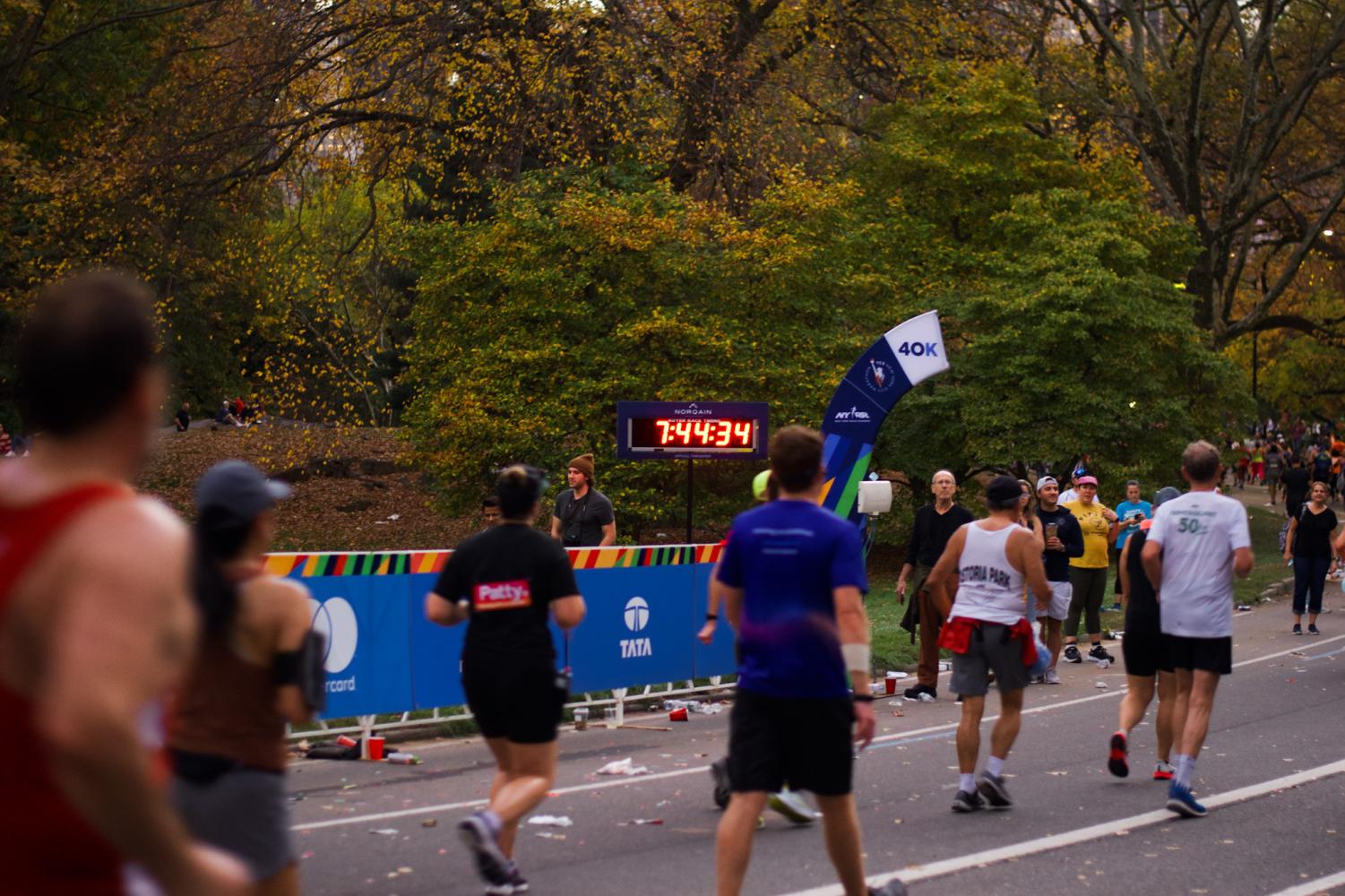 Runners wear blue, black and white shirts, and behind them is a clock that reads “7:44:34” in red text. Next to this clock, an arched sign reads “40 K”.