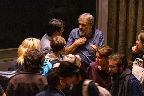 oward the top of the image, an older, bearded Slovenian man speaks to an audience member while gesturing with his hands. Other attendees are out of focus in the foreground.