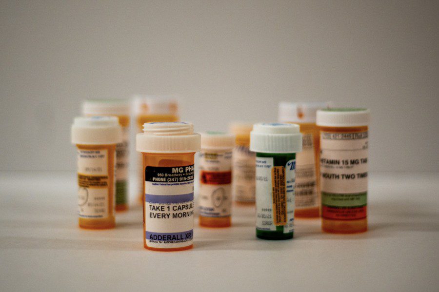 Nine cylindrical medicine canisters laid across a white table. The canister in the center is labeled “ADDERALL X.R.,” and has the instructions “TAKE 1 CAPSULE EVERY MORNING” printed on it.