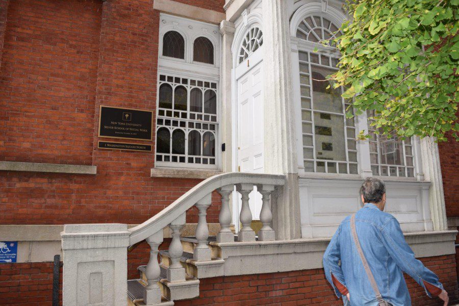 The facade of a brick building. A small plaque reads: “New York University Silver School of Social Work.” To the right, a gray-haired man wearing a jean jacket walks by.