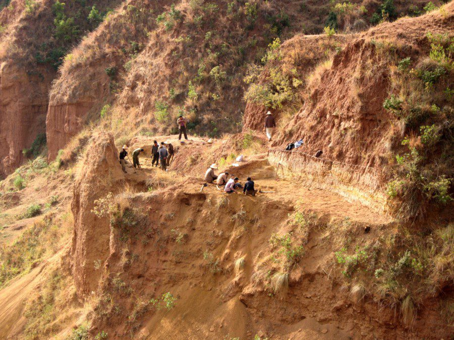 A landscape photograph of a sandy cliffside that levels off and features a group of around a dozen people at the top. The people are excavating. They have shovels with them and are wearing long sleeve shirts and pants, and some have hats.