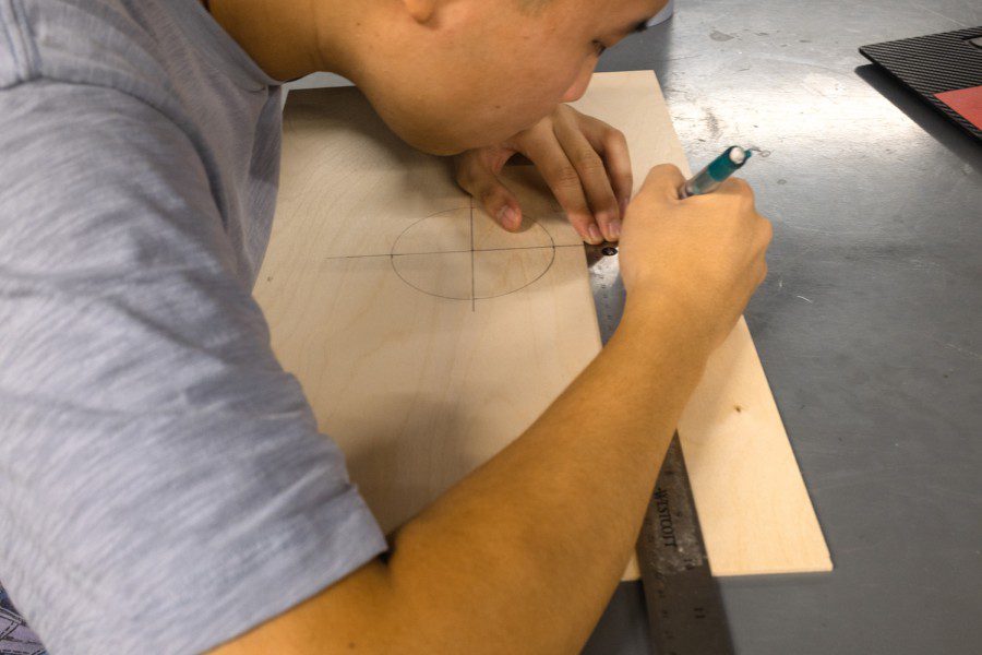 Alex Xu wears a gray T-shirt and outlines the shape of the rocket fins with a pencil and a ruler on a piece of plywood.