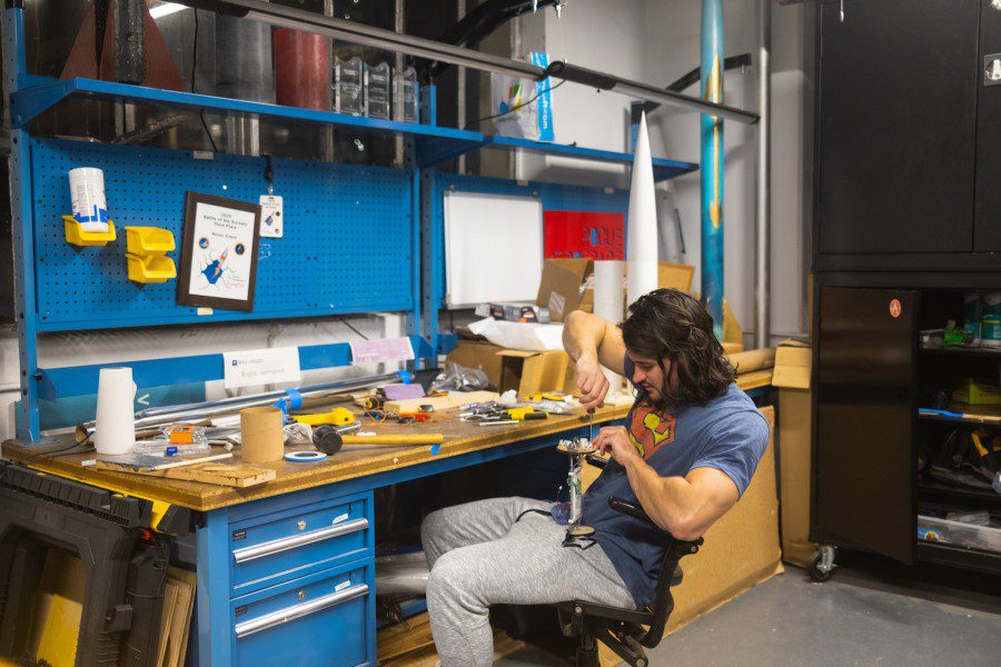 Alex Miller wearing a blue-and-red Superman T-shirt while screwing electronic sensors onto a piece of wood at his workshop.