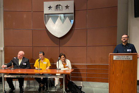 Three people sit behind a desk with three microphones in front of them while another person stands behind a podium with a plaque labeled “College of Arts & Science.” From the left: a man with a yellow shirt and black suit jacket; a woman with a bright yellow shirt; a woman with an orange shirt and white coat. The man behind the podium is wearing glasses and a dark blue shirt.