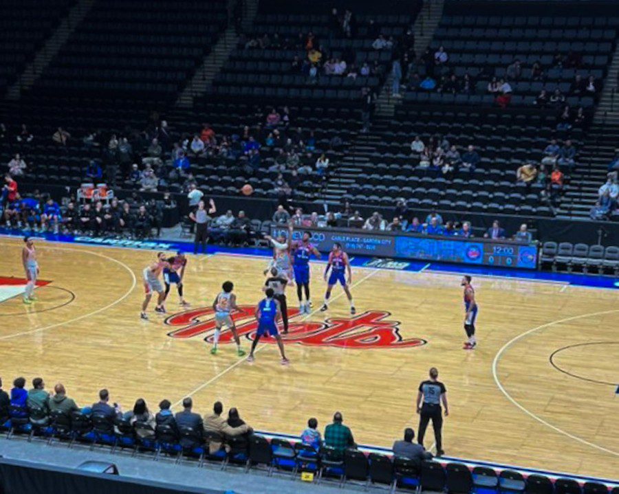 A photo of a basketball court with rows of seats in the background, athletes in blue and white uniforms playing, and a jumbotron screen hanging above.
