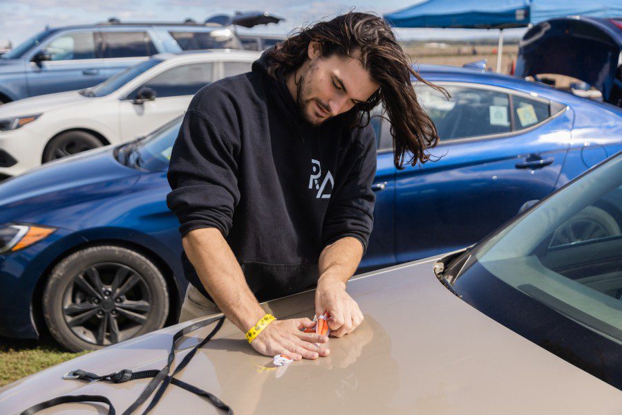 Alex Miller folds a drogue for his rocket, which is sitting on the hood of a car in a parking lot.