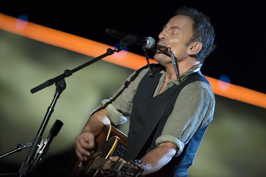 Bruce Sprinsteen, wearing a green shirt and a black-striped vest, plays a guitar and harmonica against a green, orange and black background.