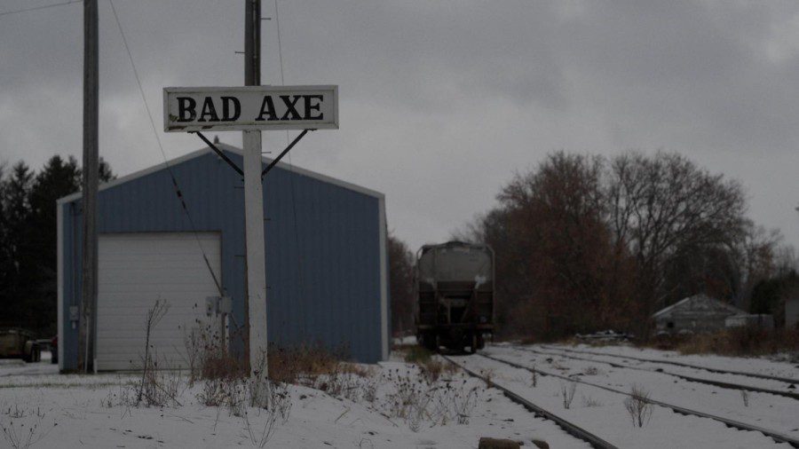 A desaturated image depicting a light blue warehouse next to train tracks, with a black train approaching it. In front is a white sign with black text that reads “BAD AXE.” There is snow on the ground.