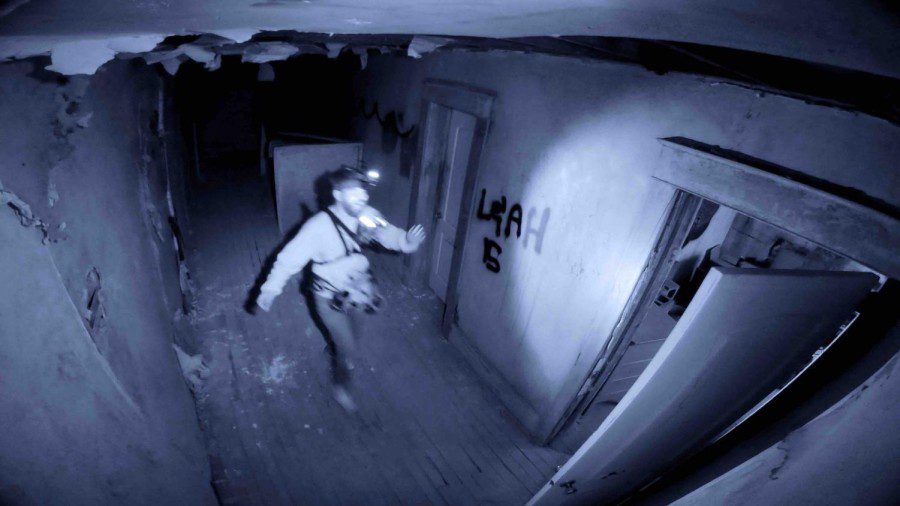 A still from the movie “Deadstream” in which a white middle-aged man runs across a dark hallway. The man is wearing a GoPro camera on his forehead.
