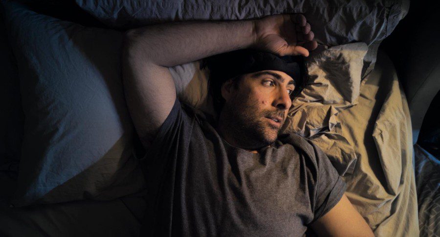 A bearded man wearing a gray T-shirt and a black hat lies on a bed with gray sheets. His arm rests above his head.
