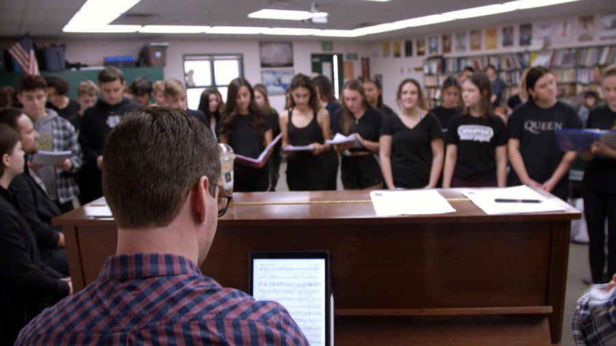 A man wearing a shirt with blue-and-red grid patterns playing a piano. A group of students stand behind the piano singing and wearing black clothes. They are inside a classroom.