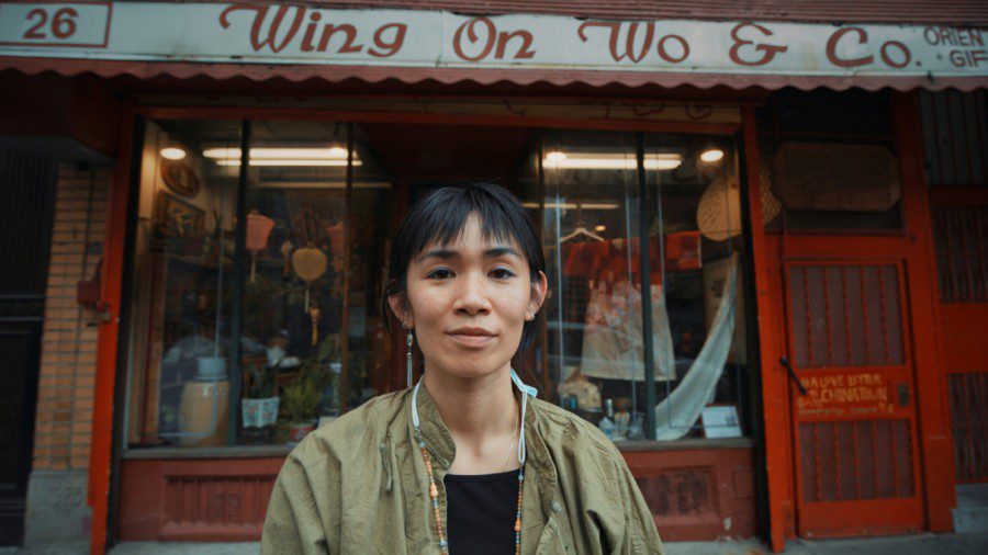 A portrait of an Asian woman in front of a store. The banner on top of the window reads “Wing On Wo & Co.” The woman has black short hair with bangs. She wears a beaded necklace, a black shirt and a military green jacket.