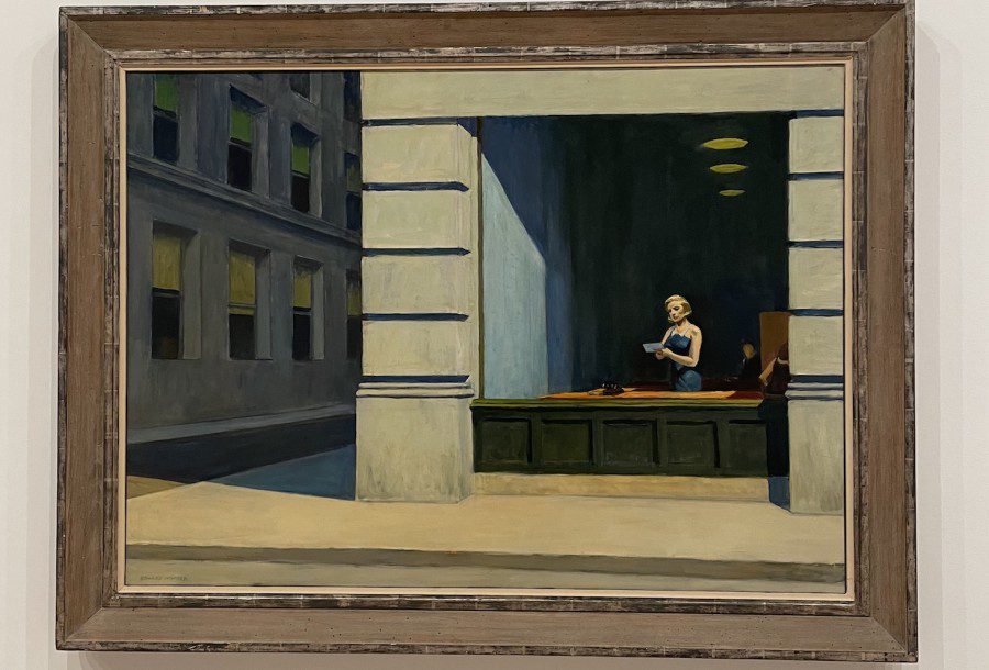 The painting “New York Office” by Edward Hopper. A young woman stands next to a large window. She wears a blue dress and holds a letter. The room has light blue walls and warm lighting, and the building has a white marble facade. The sun shines diagonally into the scene, casting a shadow on the adjacent street.