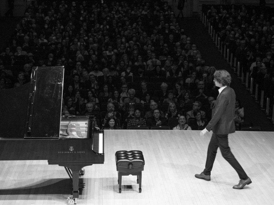Pianist Lucas Debargue wearing a black suit and white shirt walking toward a Steinway & Sons grand piano on stage with a large audience sitting in the background. The photo is black and white.