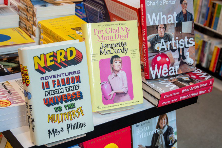 The cover of the book “I’m Glad My Mom Died” is centered in frame. Pink text against a light yellow background reads “I’m Glad My Mom Died.” The author’s name, “Jennette McCurdy,” is pink and outlined in black. At the center of the cover is a portrait of actor Jennette McCurdy, whose hair is tied up into a ponytail. She wears a pink dress and holds a pink urn against a pink background.