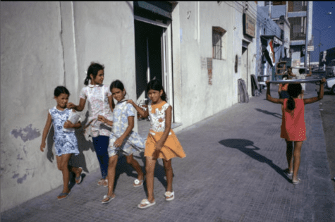 Four girls wearing colorful dresses walk down a street, while another child holding a metal plate over her head walks in the opposite direction.