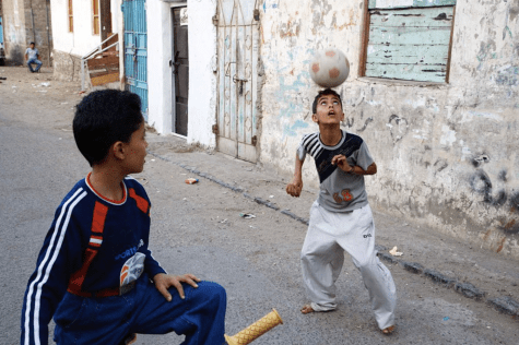 Two boys play with a soccer ball. On the left is a boy wearing a blue jersey and jeans on a bicycle. He is looking at the other boy, who is wearing a gray T-shirt and pants while reaching for the ball with his head.