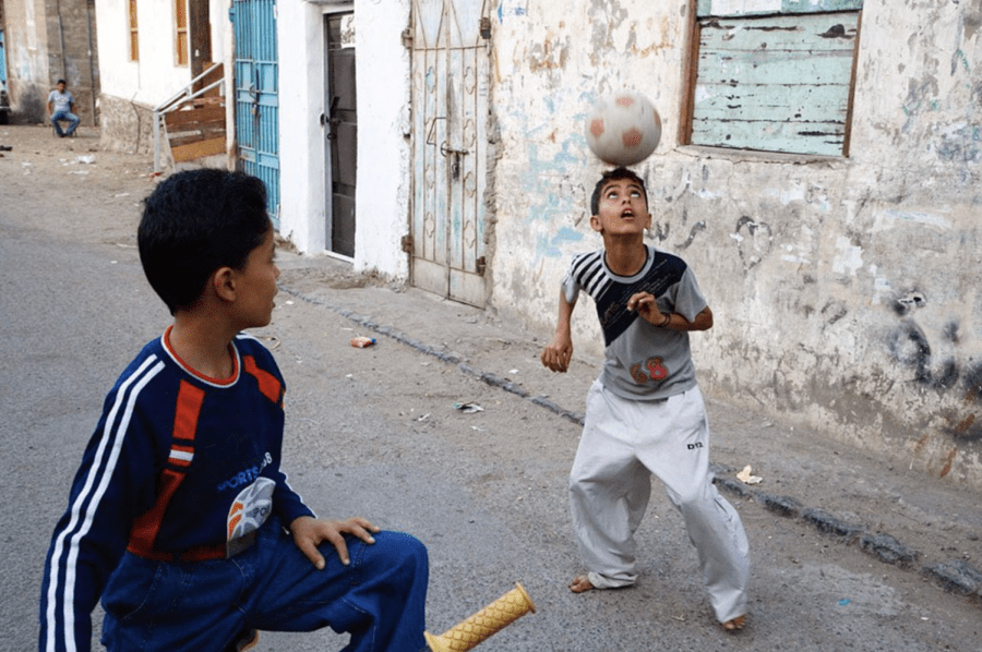 Two+boys+play+with+a+soccer+ball.+On+the+left+is+a+boy+wearing+a+blue+jersey+and+jeans+on+a+bicycle.+He+is+looking+at+the+other+boy%2C+who+is+wearing+a+gray+T-shirt+and+pants+while+reaching+for+the+ball+with+his+head.