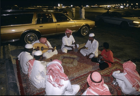 Nine men sit in a circle by the side of a street at night. A brown car is parked behind them and a white car is passing by. Eight of them are wearing white Arabic thawb; one is wearing a red T-shirt and black pants. The two people sitting closest to the parked car are holding musical instruments.