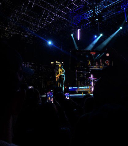 Photo of a concert. On stage is a man wearing all-black playing a guitar. The stage is flooded with various colored lights.