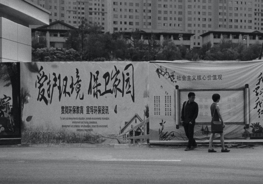 A black and white still image from a film depicting a man and a woman walking past each other against a sign filled with propaganda posters in Mandarin.