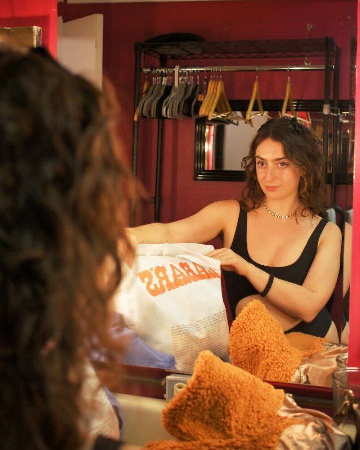 Actress Sophie Zucker wearing a black vest looking into a mirror inside a dressing room with red walls and a clothing rack with many hangers on it.