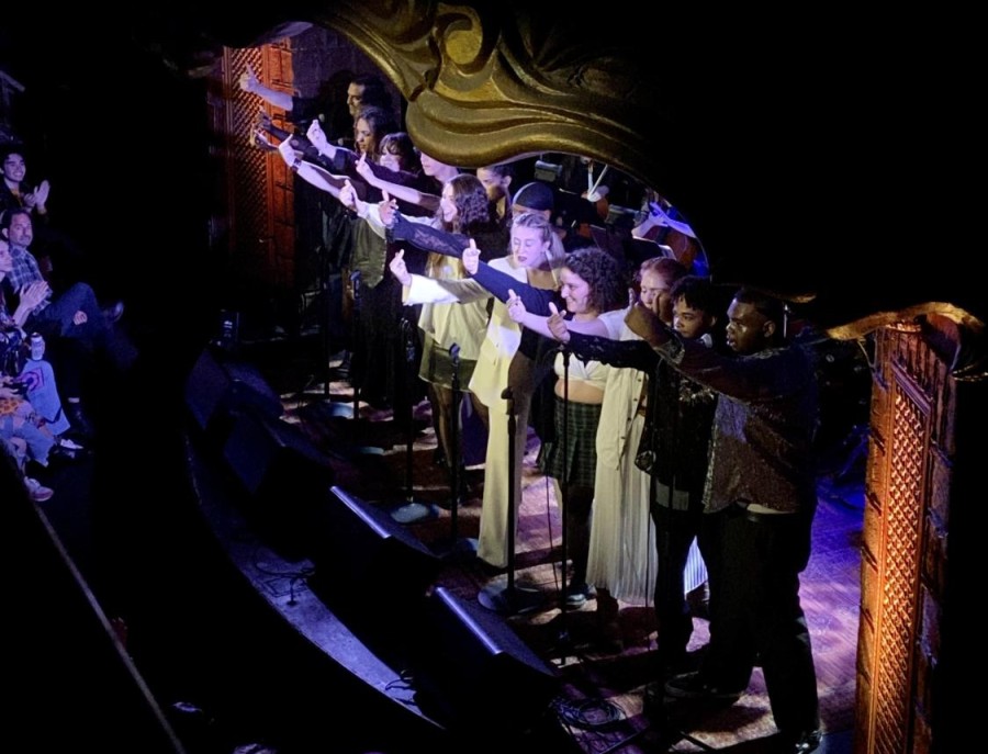 A line of performers dressed in black and yellow stand on stage, making hand gestures toward the audience.