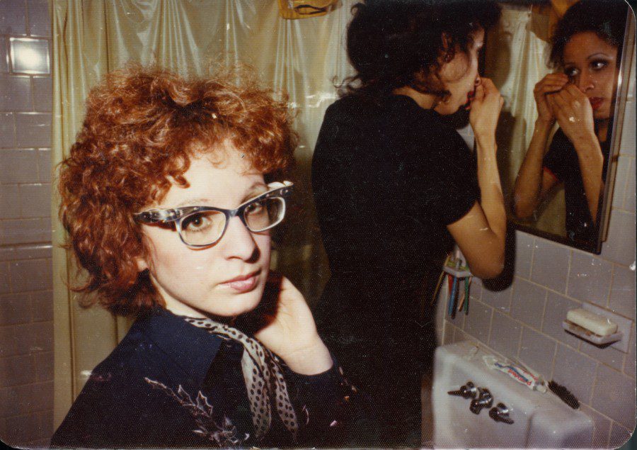 A+photograph+of+two+girls+in+a+bathroom+near+the+sink+and+mirror.+One+girl+has+curly+red+hair%2C+black+rimmed+glasses%2C+black+clothes+and+is+looking+into+the+camera.+The+second+girl+is+wearing+black+clothes%2C+short+black+hair+and+is+looking+into+the+mirror+fixing+her+eyelashes.